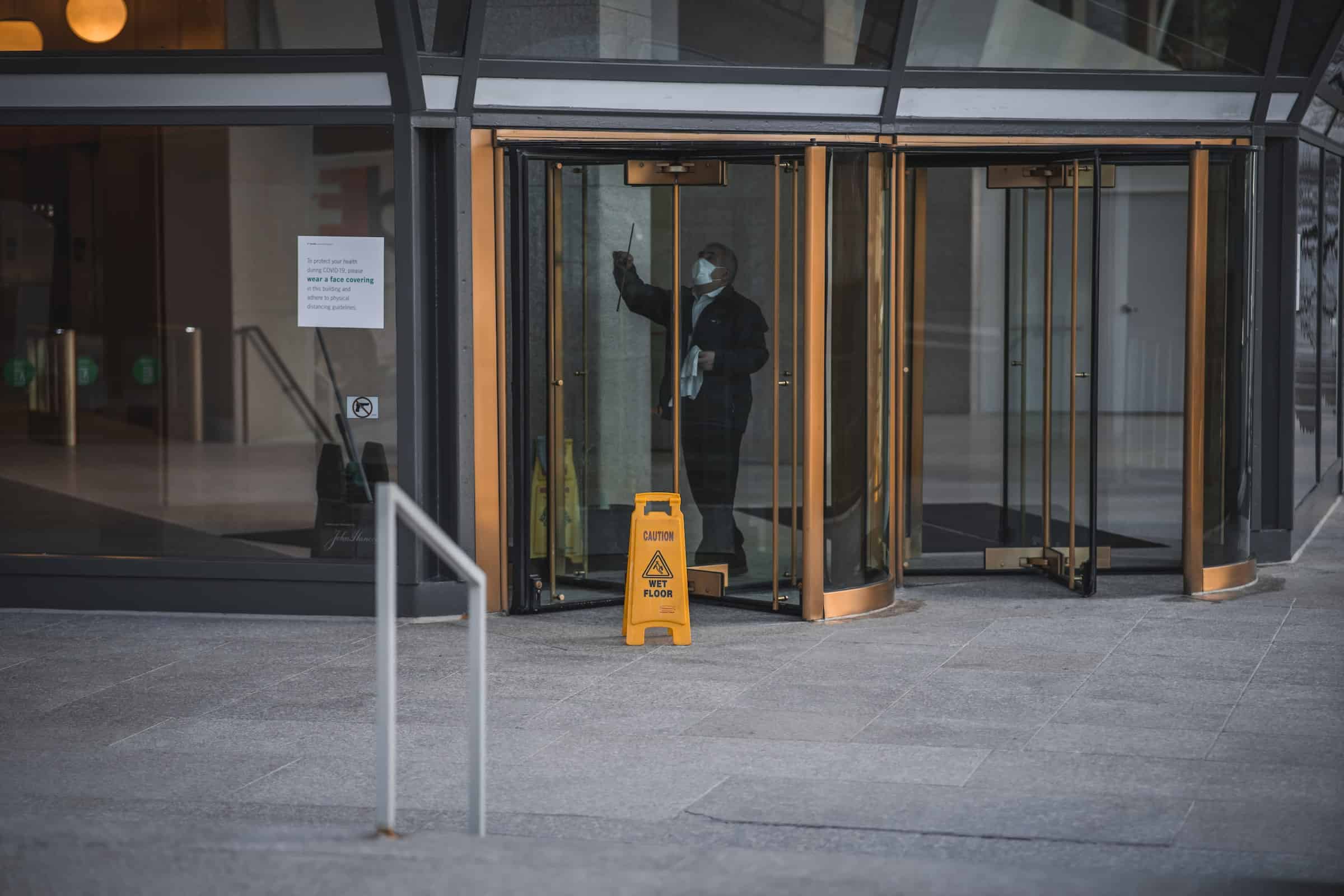 Masked day porter cleaning and disinfecting entrance to office building